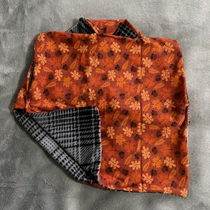 May include: A reversible blanket featuring a vibrant orange side with a repeating pattern of brown acorns and various autumn leaves, and a contrasting side with a black and white houndstooth check pattern, showcasing a soft fabric texture.