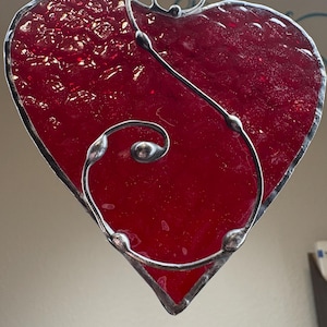 May include: A handcrafted, red stained glass heart with a textured surface. The heart is outlined with a silver-colored metal and features a decorative, swirling metal accent. A small loop at the top allows for hanging.