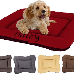 May include: Red dog bed with a black embroidered name "Daizy" on the front. The bed has a raised edge and a soft, plush surface. The bed is shown with a small dog lying on it. The image also shows four other dog beds in different colors: brown, gray, beige, and brown.