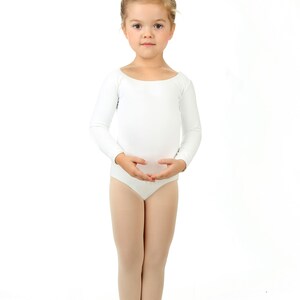 May include: A young girl wearing a white leotard and ballet shoes stands with her arms crossed in front of her. She has her hair pulled back in a bun.