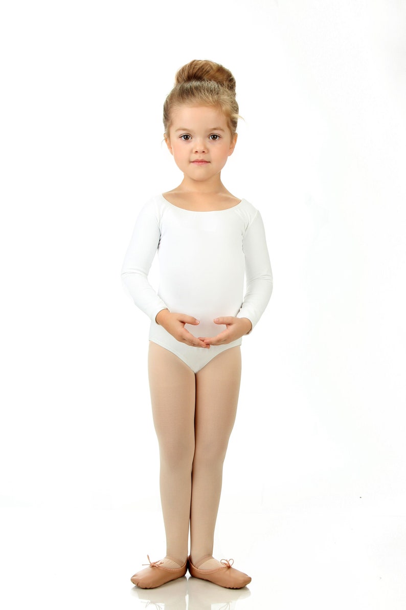 May include: A young girl wearing a white leotard and ballet shoes stands with her arms crossed in front of her. She has her hair pulled back in a bun.