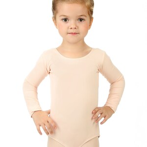 May include: A young girl wearing a light pink leotard with long sleeves. She has her hair pulled back in a bun and is standing with her hands on her hips.