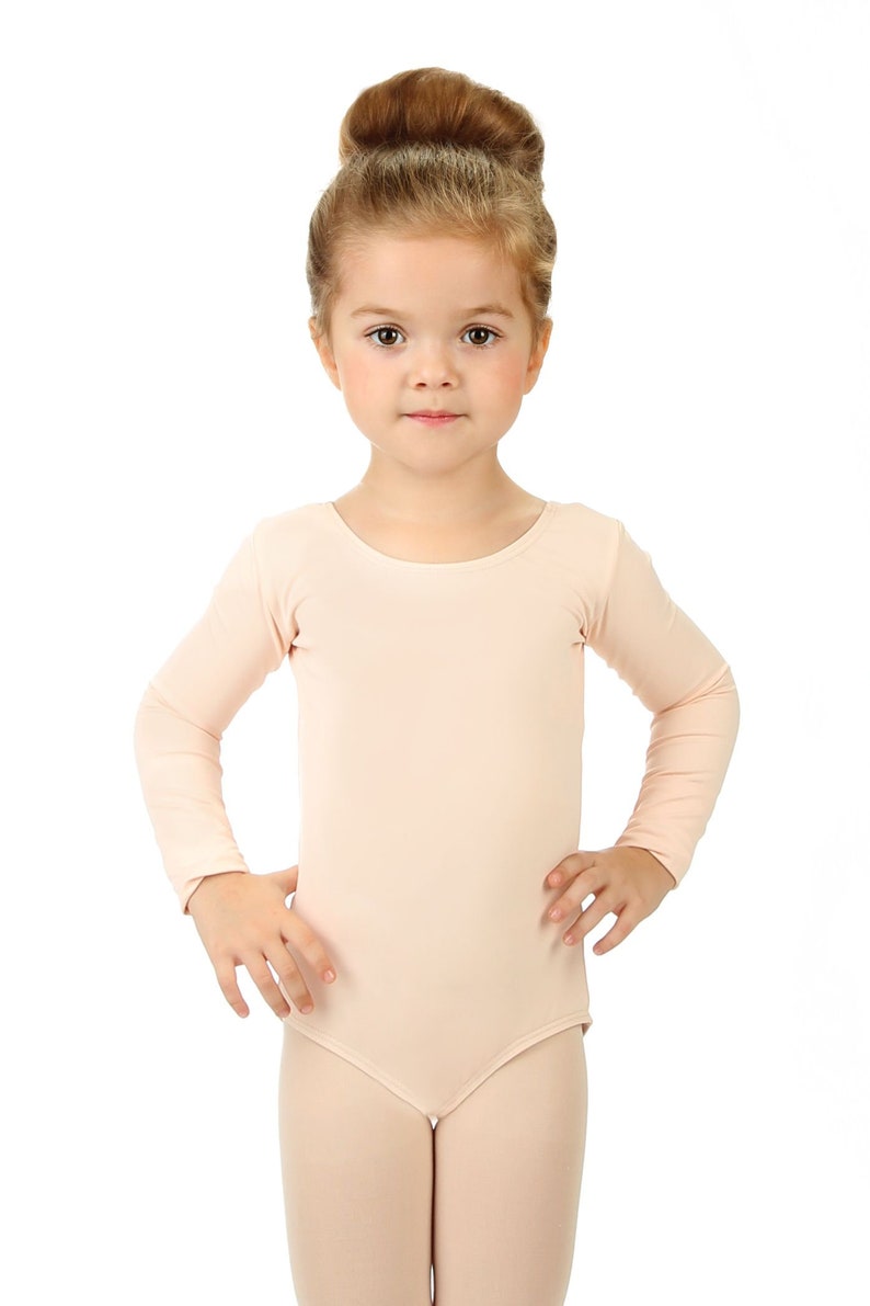 May include: A young girl wearing a light pink leotard with long sleeves. She has her hair pulled back in a bun and is standing with her hands on her hips.