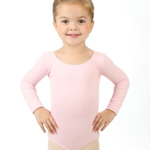 May include: A young girl wearing a pink long-sleeved leotard, smiling at the camera.