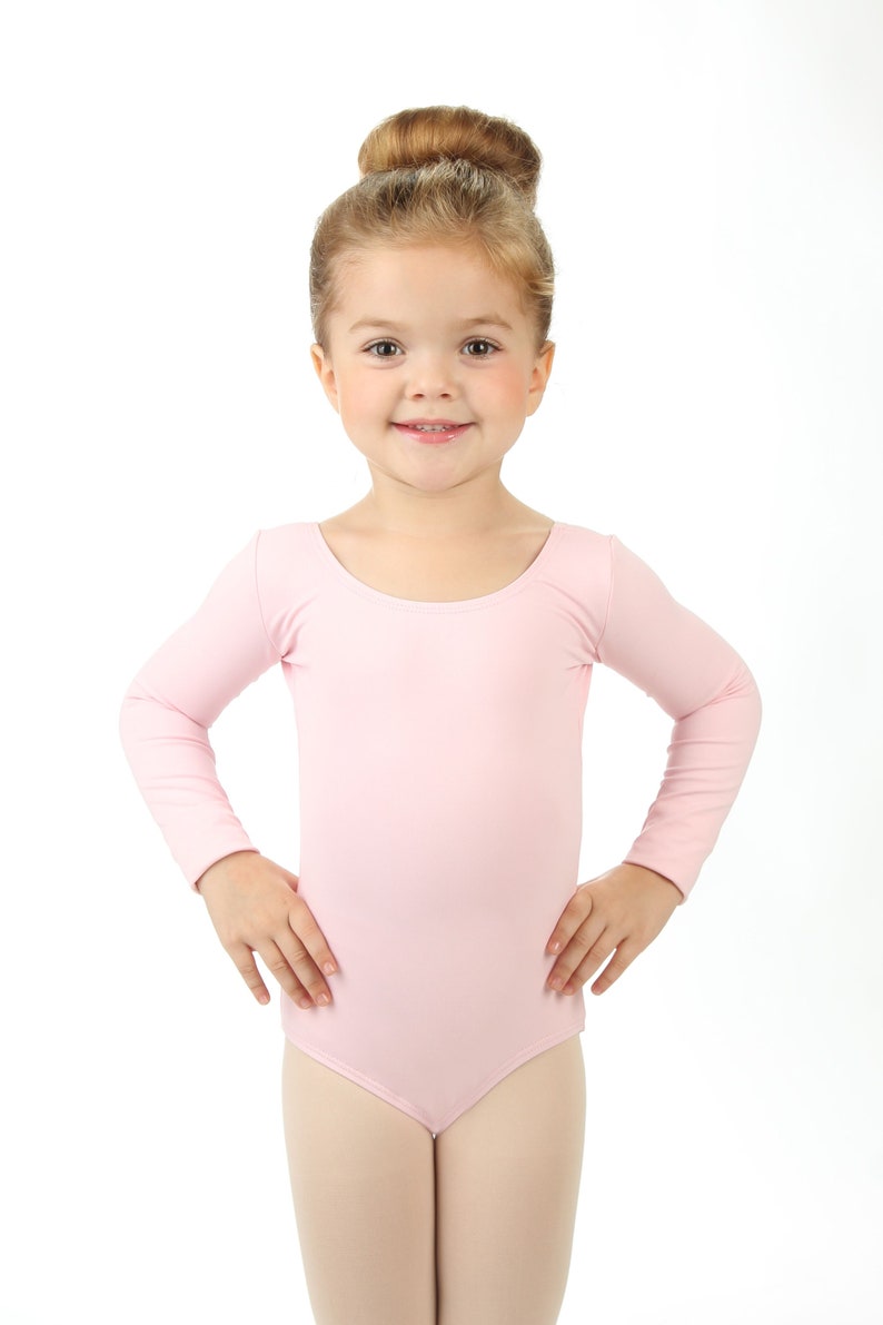 May include: A young girl wearing a pink long-sleeved leotard, smiling at the camera.