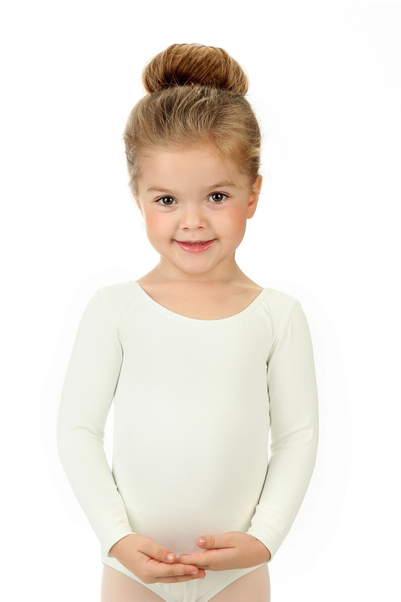 May include: A young girl with blonde hair in a bun is wearing a white long-sleeved leotard. She is smiling and looking at the camera.