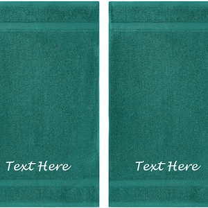 以下が含まれることがあります： テクスチャ加工された表面の、ターコイズ色のバスタオル2枚。各タオルは、上部と下部に濃いターコイズ色のボーダーがあります。「Text Here」の文字が、各タオルの下部に白い筆記体で書かれています。
