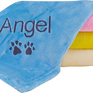 May include: A blue fleece blanket with the name "Angel" and two paw prints embroidered on it. The blanket is folded with a pink, yellow, and cream blanket underneath.