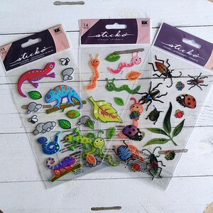 May include: Three packs of "sticko" brand dimensional stickers. The stickers feature colorful insects, reptiles, and leaves. The packs include 14 or 15 pieces each. The stickers are on a white wooden surface.