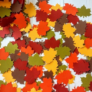 May include: A collection of paper maple leaves in various autumn colors, including red, orange, yellow, brown, and green, scattered on a white wooden surface.