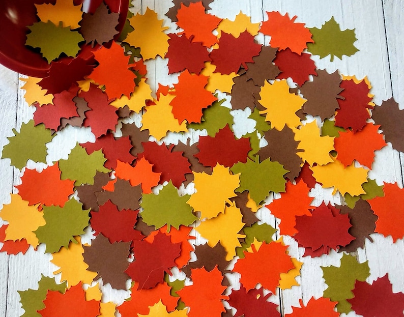 May include: A collection of paper maple leaves in various autumn colors, including red, orange, yellow, brown, and green, scattered on a white wooden surface.