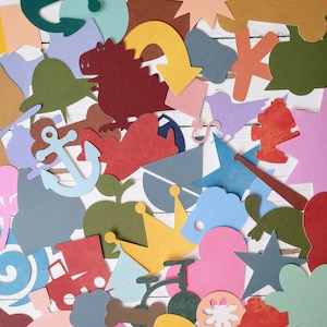 May include: A collection of colorful paper cutouts in various shapes, including a crown, an anchor, a star, a heart, a fish, a flower, and a train. The cutouts are arranged on a white wooden surface.