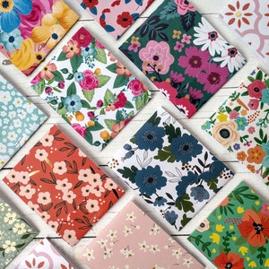 May include: A collection of 12 small, square notebooks with floral patterns. The notebooks have a variety of floral designs, including blue, pink, red, and green flowers. The notebooks are arranged in a diagonal pattern on a white wooden surface.