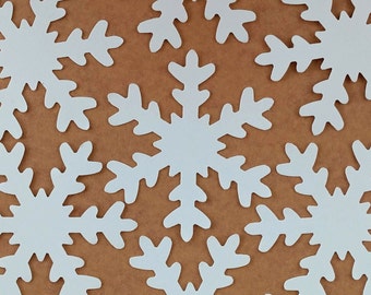 May include: White snowflake cutouts arranged on a brown background. The snowflakes have intricate, symmetrical designs, creating a winter or holiday theme. The image is a flat lay, with the snowflakes evenly distributed.