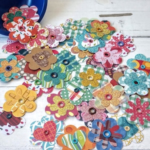 May include: A collection of colorful paper flower embellishments with various patterns and textures. The flowers feature glitter, buttons, and patterned paper in shades of red, blue, green, yellow, and brown. A blue bowl is visible in the background.