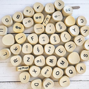 May include: A collection of beige wooden tiles with various letters, numbers, and symbols printed on them. The tiles are arranged in a circular pattern on a white wooden surface.