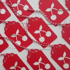 May include: Red gift tags featuring a white cherry design and white twine. The tags have a scalloped edge and are ideal for adding a festive touch to gifts.