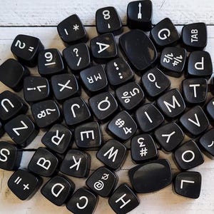 May include: A collection of black, square-shaped keyboard keys with white lettering and symbols. The keys include letters, numbers, and special characters. The keys are scattered on a light-colored wooden surface.