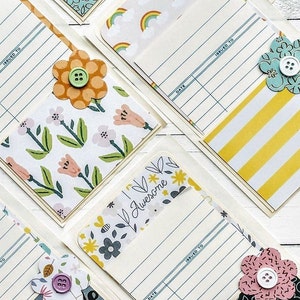 May include: Six handmade cards with a vintage library card theme. Each card features a different patterned paper with a flower embellishment. The cards have a blank space for writing a message and a space for the date and to whom the card is issued.