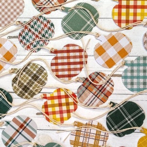 May include: A collection of 16 fall-themed pumpkin-shaped paper cutouts with various plaid patterns in shades of green, brown, orange, and white. The cutouts are strung together with twine.