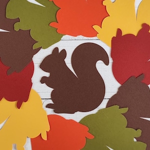 May include: A collection of paper squirrel cutouts in various autumn colors including dark brown, olive green, bright orange, yellow, and deep red. The shapes are arranged in a cluster, featuring a central dark brown squirrel silhouette.