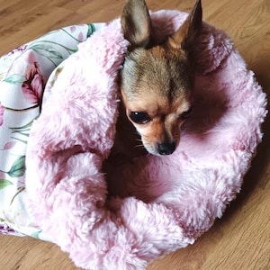 Puede incluir: Un pequeño perro marrón y blanco está acurrucado en una cama para perros rosa y blanca con un patrón floral. La cama está hecha de tela suave y de felpa.
