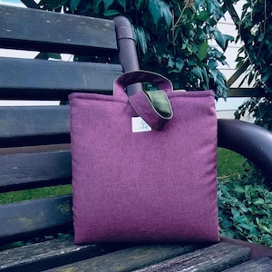 Puede incluir: Un bolso tote de color ciruela con una tela texturizada, con un asa y ribete verdes. El bolso está sobre un banco de madera con un fondo natural de follaje verde. Una pequeña etiqueta blanca es visible en la parte delantera del bolso.