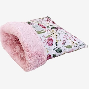 Puede incluir: Una cama para mascotas rosa con un interior de piel sintética afelpada y un exterior con estampado floral. El estampado floral presenta flores rosas y moradas con hojas verdes sobre un fondo blanco. La cama está diseñada para la comodidad de las mascotas pequeñas.