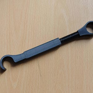 May include: A black, 3D-printed tool featuring a central rectangular handle, a threaded adjustment section, and two distinct circular ends. One end is a closed ring, while the other end is an open, C-shaped hook designed for gripping or turning.