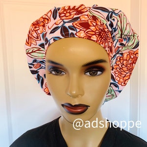 May include: A floral patterned scrub cap in shades of pink, orange, brown, and teal. The cap features a large, colorful floral design. The text "@adshoppe" is visible on the lower right.