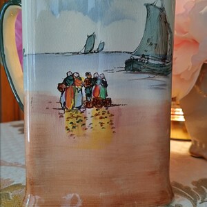May include: A ceramic pitcher with a coastal scene painted on the front. The artwork depicts figures on a beach with sailboats in the distance. The pitcher has a green handle and rim, with a peach-colored base.