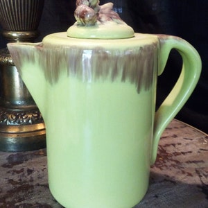 May include: A green ceramic coffee pot with a brown glaze and an acorn-shaped lid. The lid is decorated with three acorns.