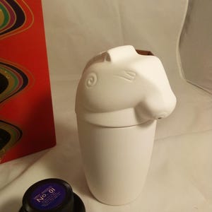 May include: A white ceramic container with a horse head design on top. The container is next to a bottle of ink and a red box with a colorful design.