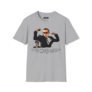 May include: A light gray t-shirt featuring a graphic of a man flexing his biceps, wearing sunglasses and a suit. The text "BIG FLEX COMPLEX NAPOLEON" is below the image. The shirt has a crew neck and short sleeves.
