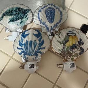 Peut inclure: Quatre veilleuses décoratives dotées d'abat-jours en coquillages naturels. Les coquillages sont ornés d'images d'une tortue de mer, d'un blason bleu, d'un crabe bleu et d'un oiseau jaune et bleu perché sur une branche. Chaque base est blanche.