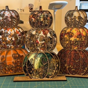 May include: Three stacked pumpkin decorations made of laser-cut wood with intricate floral and leaf outlines. Each pumpkin features a different colorful patterned insert, including orange plaid, autumn leaves, and green foliage designs on flat bases.