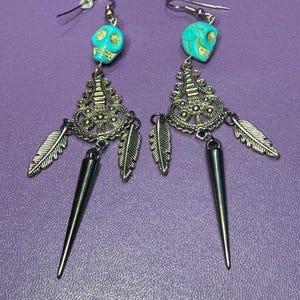 May include: A pair of dangle earrings with turquoise skull charms, silver filigree, and feather accents. The earrings have a silver spike at the bottom.