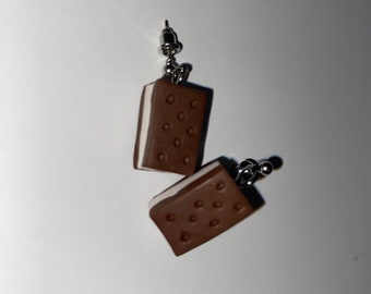 May include: A pair of earrings shaped like ice cream sandwiches. The earrings are made of brown and white clay and have a silver hook.