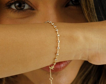 May include: A close-up of a woman wearing a gold chain necklace and a gold beaded bracelet. The woman is wearing a white sweater and has long dark hair.