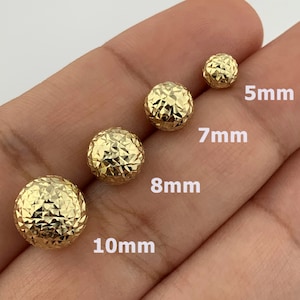 Może przedstawiać: Cztery złote, okrągłe kolczyki w różnych rozmiarach: 5 mm, 7 mm, 8 mm i 10 mm, są prezentowane na dłoni. Kolczyki mają fakturowane, diamentowe wykończenie.