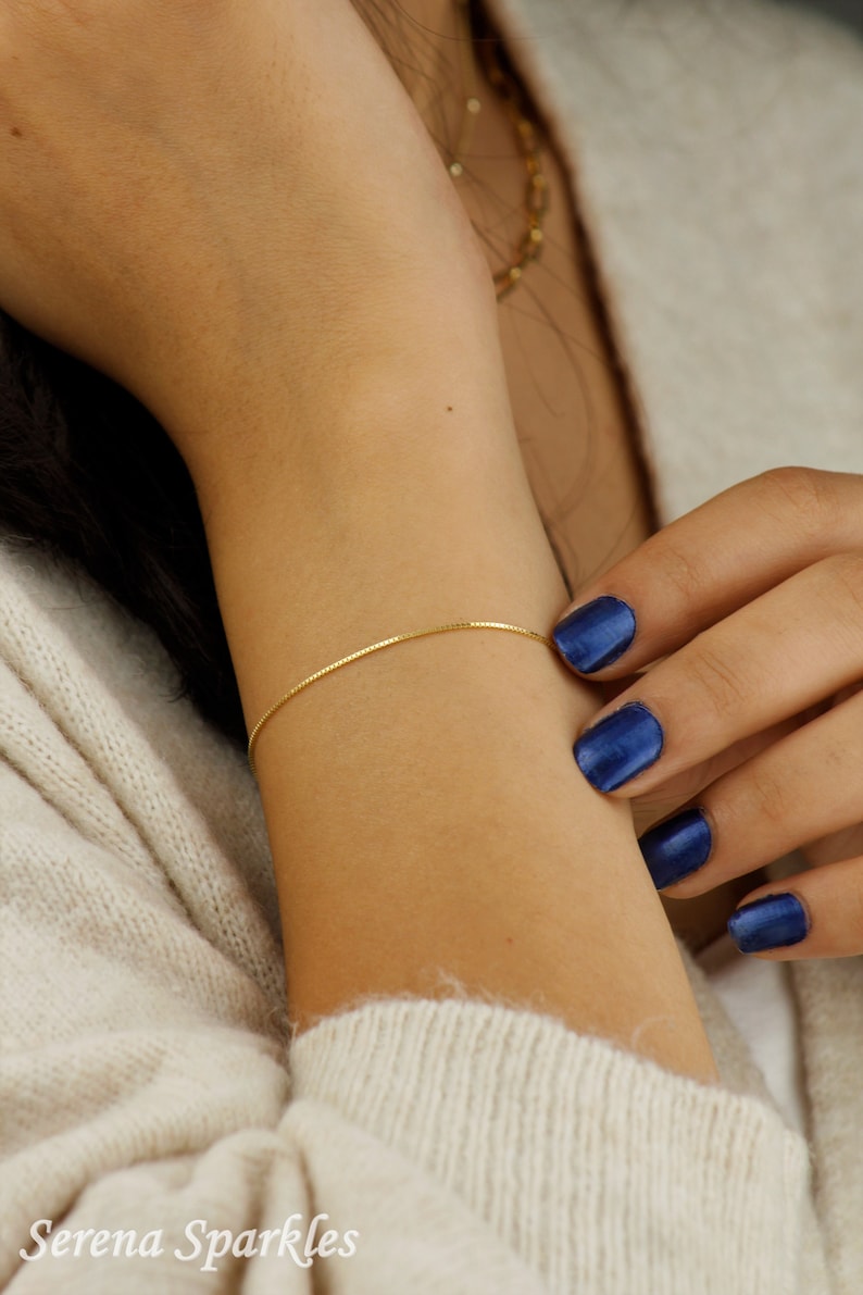 Puede incluir: Un delicado brazalete de cadena dorada en la mu&ntilde;eca de una persona. El brazalete es delgado y tiene un dise&ntilde;o de cadena de caja. Las u&ntilde;as de la persona est&aacute;n pintadas de un color azul oscuro.