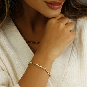 May include: A gold rope chain bracelet on a person's wrist. The bracelet is made of a twisted rope design.