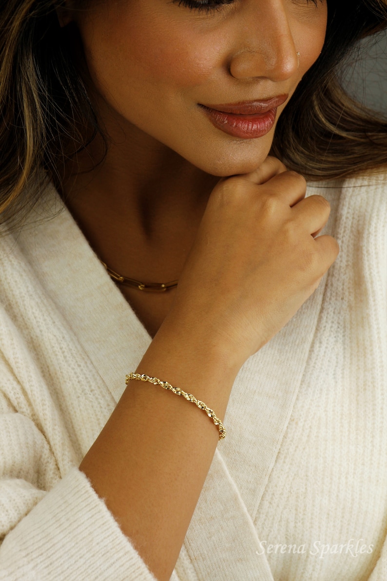 May include: A gold rope chain bracelet on a person's wrist. The bracelet is made of a twisted rope design.