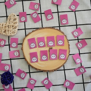 Puede incluir: Etiquetas de tela rosas con una ilustración de dumpling, dispersas alrededor de una rodaja de madera. Elementos decorativos: un corazón tejido, una bola naranja, una bola azul y un tulipán.