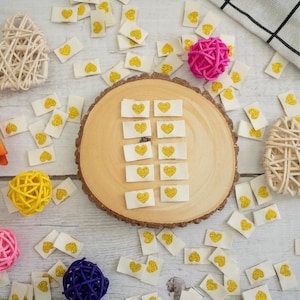 以下が含まれることがあります： 中央に円形にくり抜かれた木製の板があり、その周りを金色のハート模様の小さな白い布の正方形が囲んでいます。板の周りを、さらに多くの布の正方形、小さな白い籐製のハート、ピンク、黄色、青の毛糸玉が囲んでいます。