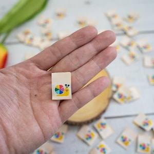 May include: A small, square, wooden tile with a colorful snail design. The snail is carrying a rainbow-colored wheel on its back. The tile is being held in a person's hand.