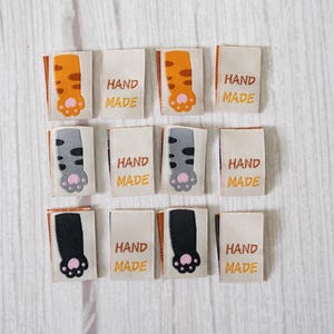 May include: Twelve rectangular fabric labels with cat paw designs. The labels feature orange, grey, and black paw prints, with the words "HAND MADE" printed in brown. The labels are arranged on a light wood-grain surface.
