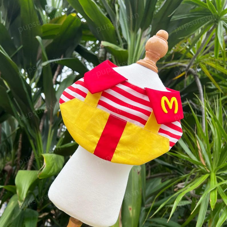 Pode incluir: Um bandana para cachorro vermelho e amarelo com listras brancas e um logotipo M amarelo. O bandana est&aacute; em um manequim branco.