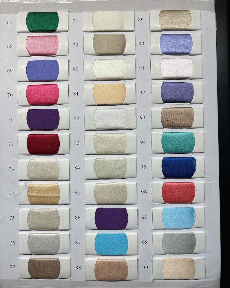 K&ouml;nnte beinhalten: Eine Stofffarbkarte mit dreiunddrei&szlig;ig rechteckigen Mustern, die in drei vertikalen Spalten angeordnet sind. Jedes Muster ist von 67 bis 99 nummeriert und zeigt eine eigene Farbe mit Satin-Finish, darunter Gr&uuml;n-, Rosa-, Blau-, Lila-, Rot-, Gold-, Silber- und Beiget&ouml;ne.