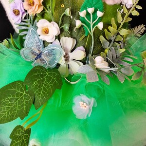 May include: A green tulle tutu with a floral embellishment featuring a blue butterfly, white flowers, and green leaves.